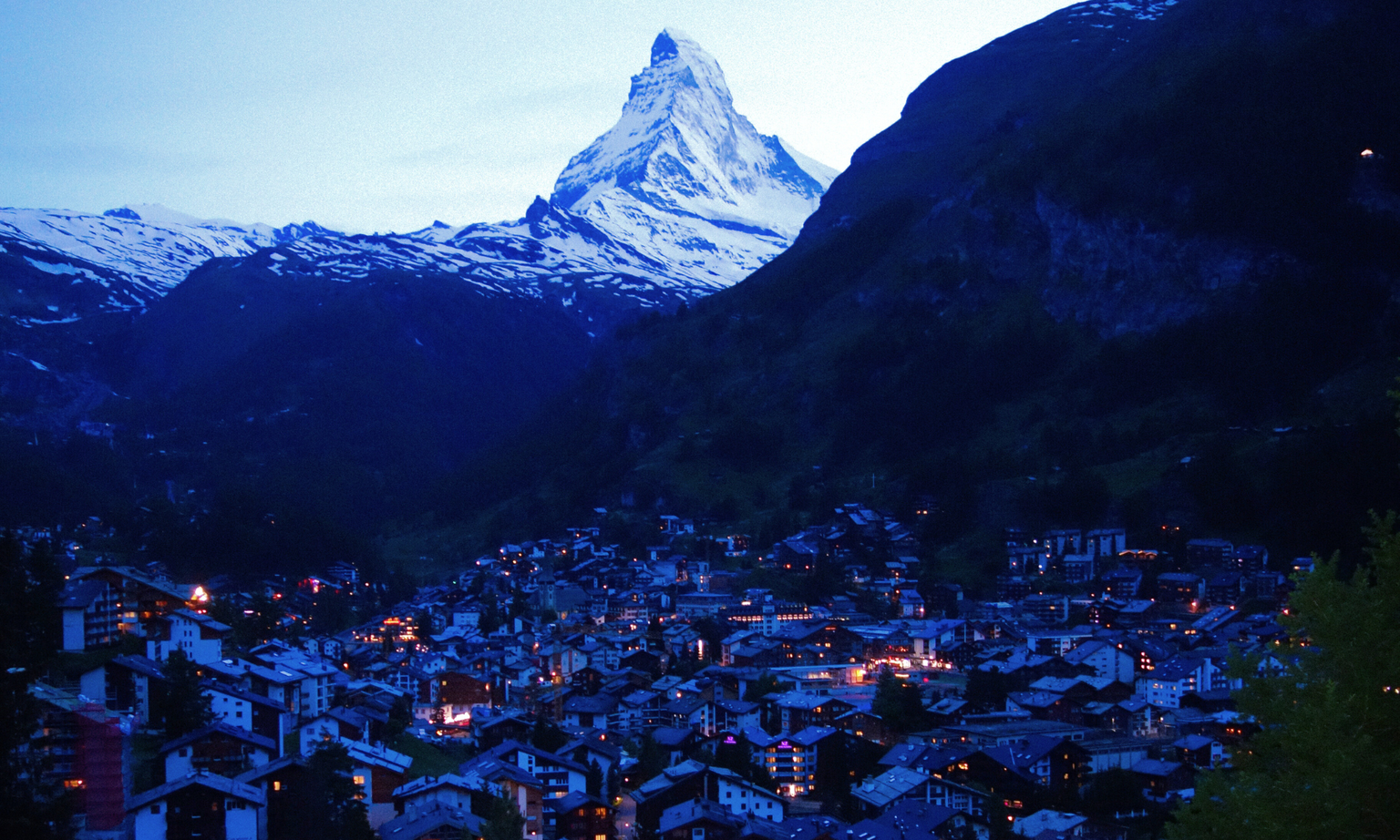 Mountain village at dusk with illuminated buildings, nestled in a valley, overlooked by a snow-capped peak against a twilight sky.
