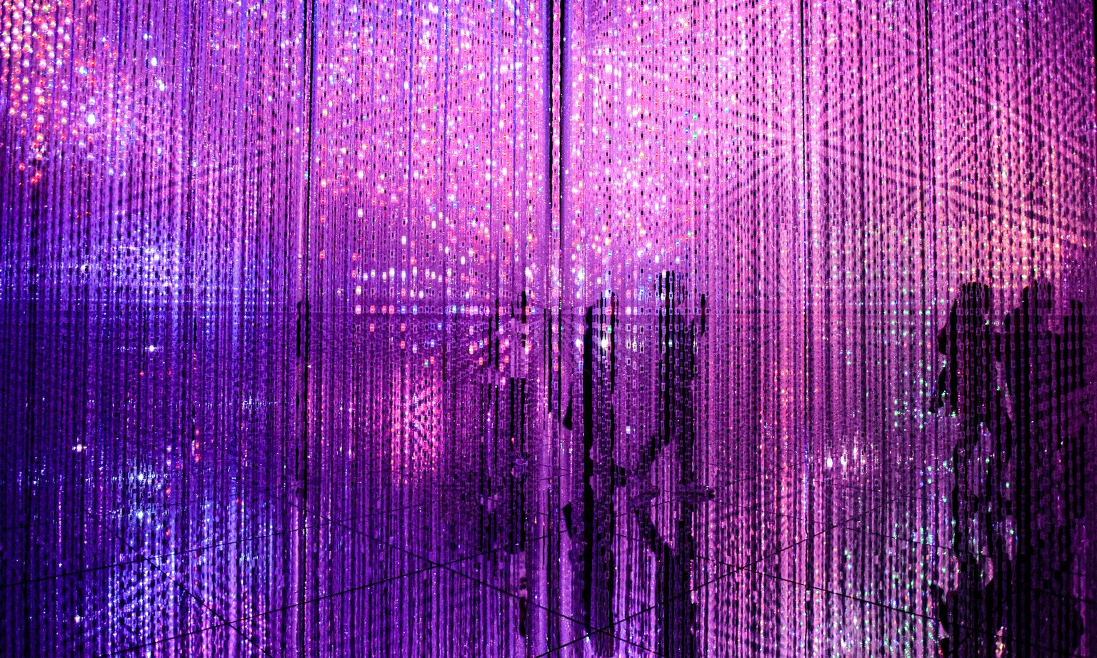 Silhouettes of people walking among LED lights hanging from the ceiling over the mirrored floor of an immersive art installation in Tokyo, Japan