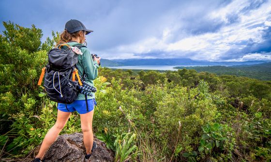 A woman in outdoor gear stands on a rock, photographing a vast, lush landscape with a lake and mountains under a cloudy sky.
