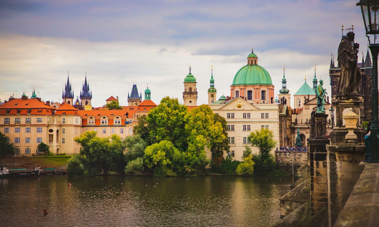 Scenic view of Prague's historic architecture, featuring colorful buildings, St. Vitus Cathedral spires, and a river under a cloudy sky.