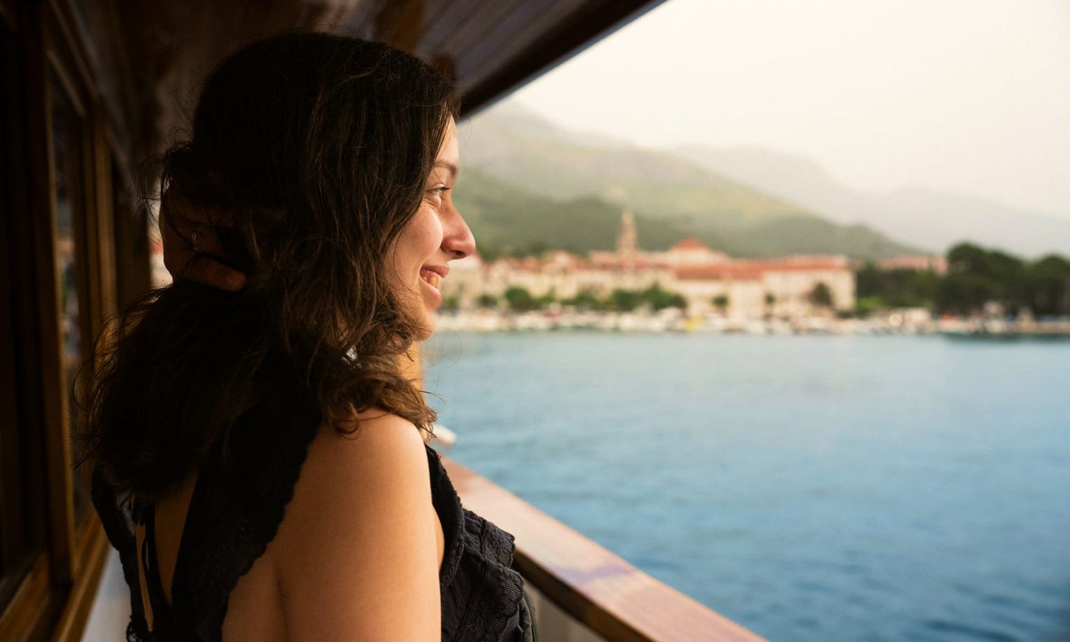 Woman gazing out from a boat at a scenic coastal town, with mountains in the background and water below.