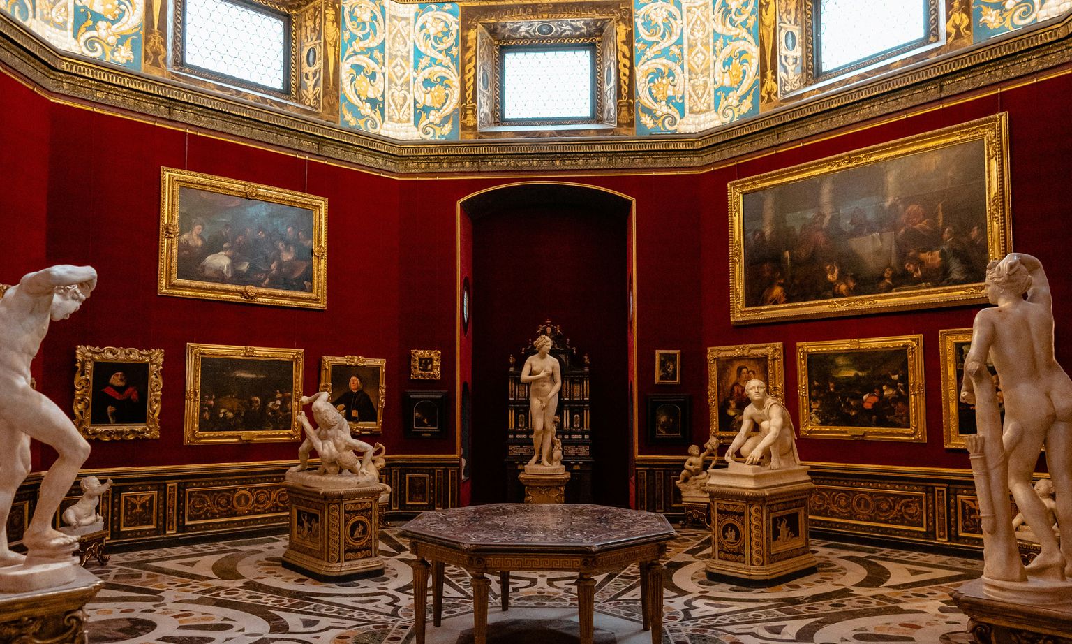 Marble statues and gold-framed paintings in a room with red walls, marble floor,  and ornate gold details at the Uffizi Gallery in Florence