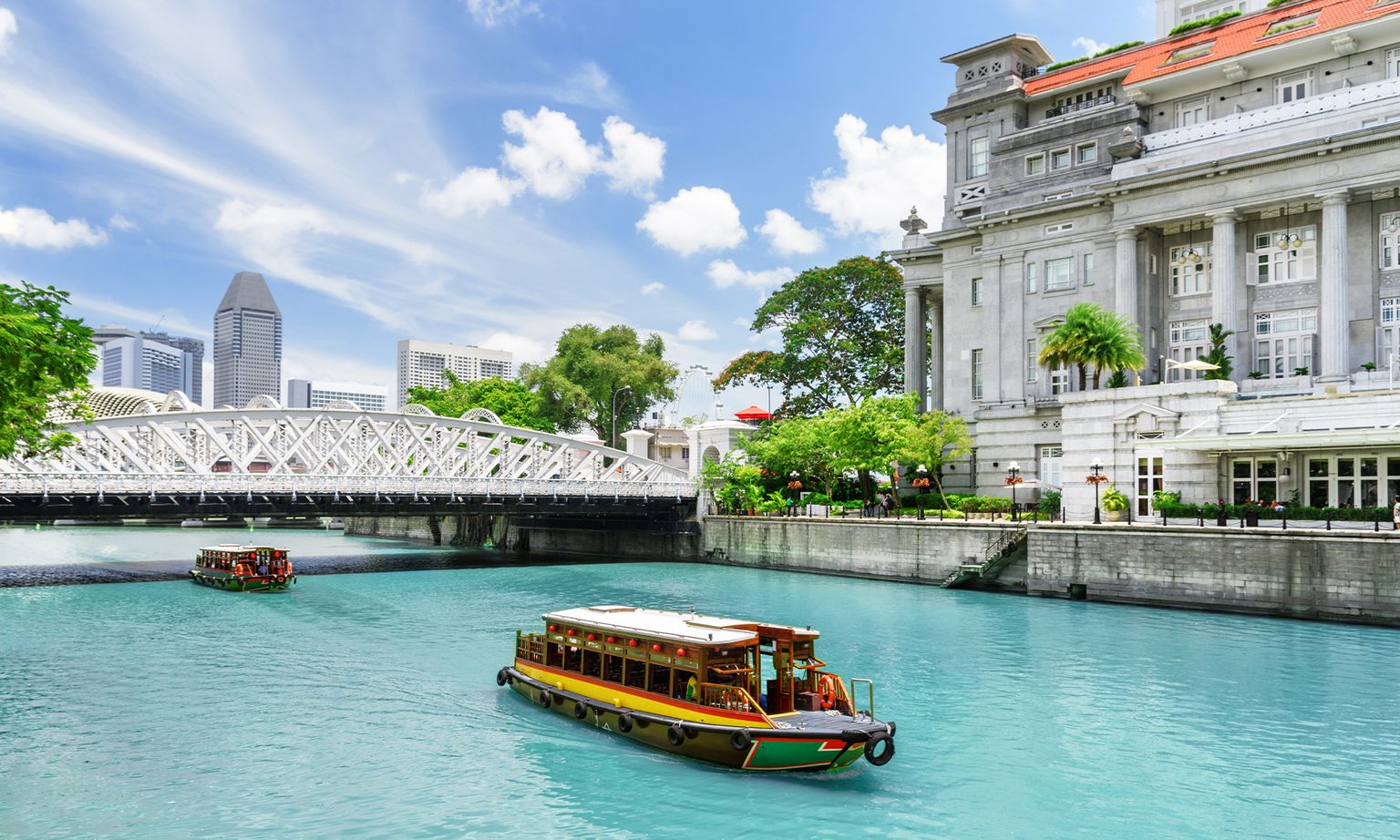 A boat on a turquoise river with a historic building and a white bridge in the background, set against a city skyline and blue sky with clouds.