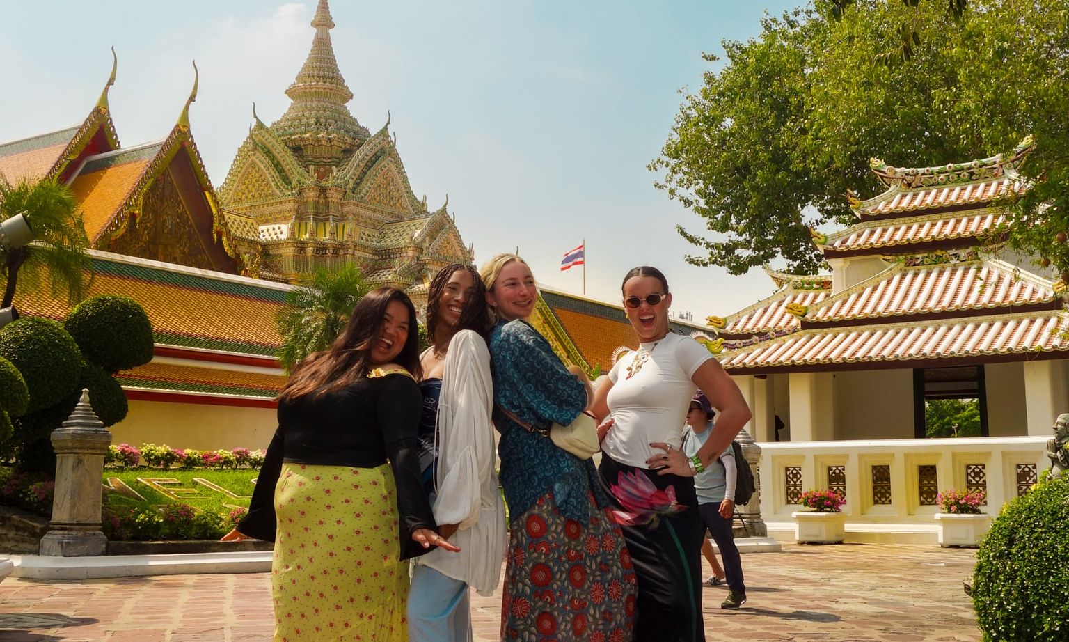 Four people posing happily in front of a traditional Thai temple with intricate rooftops and a garden. A Thai flag waves in the background.