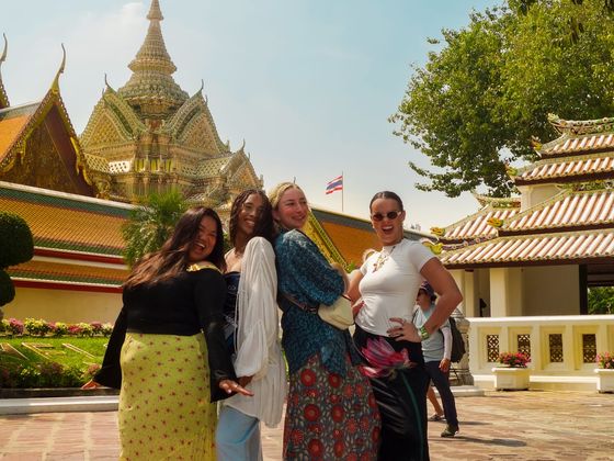 Four women smiling and posing in front of an ornate temple with a traditional Thai architectural design, under a clear blue sky.