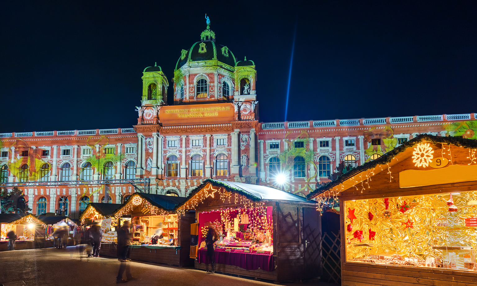 Festive Christmas market at night, with illuminated stalls and a grand, brightly lit historical building in the background.