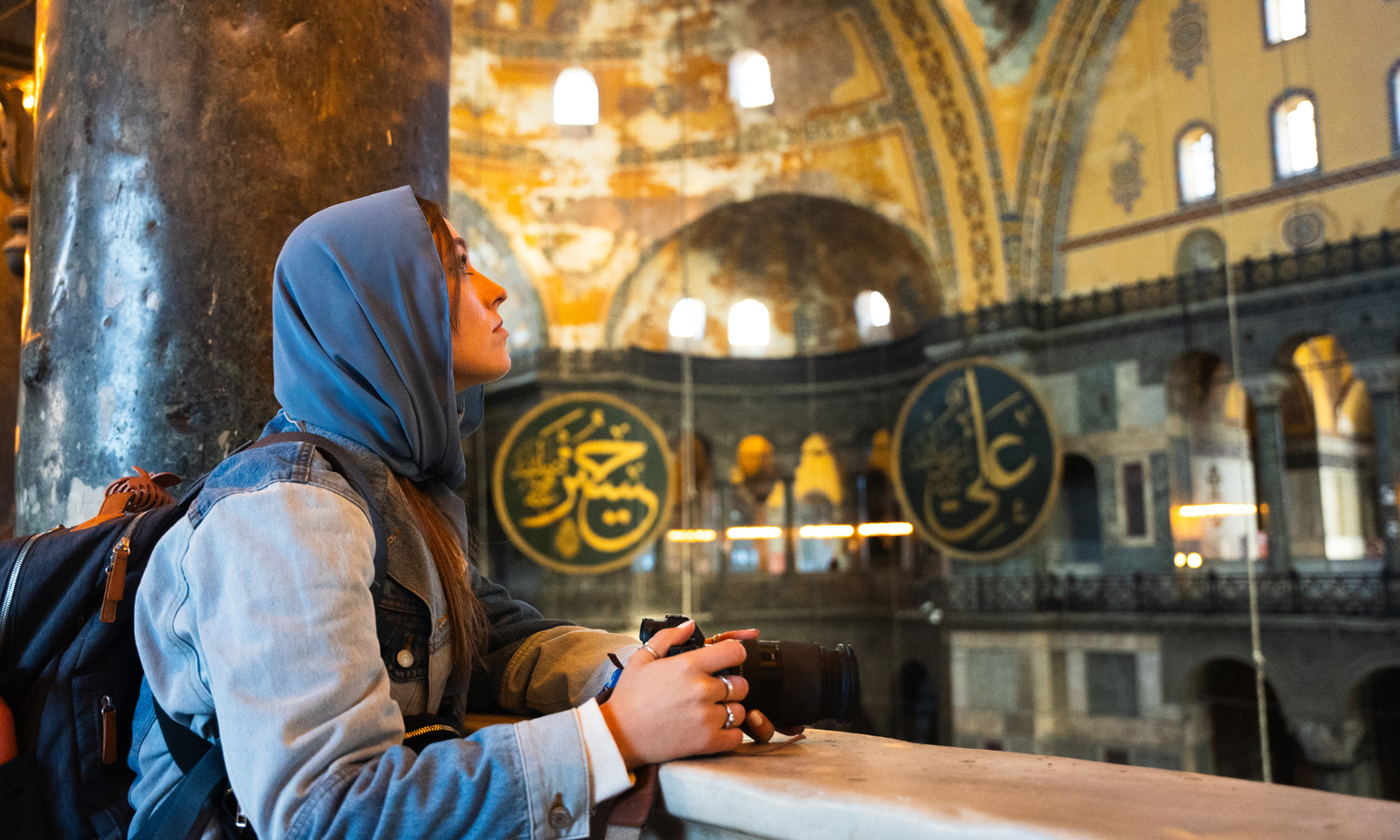 A woman in a blue headscarf and denim jacket holds a camera inside a historic building with ornate decorations and large circular calligraphy.