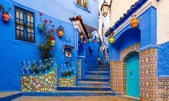 Vibrant blue alley with colorful mosaic tiles and potted plants in Chefchaouen, Morocco. Stairs lead up between uniquely decorated buildings.