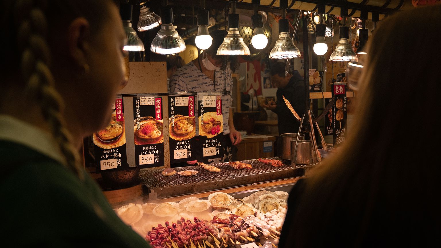 People browsing skewered food at a brightly lit street food stall with signage and lamps overhead.