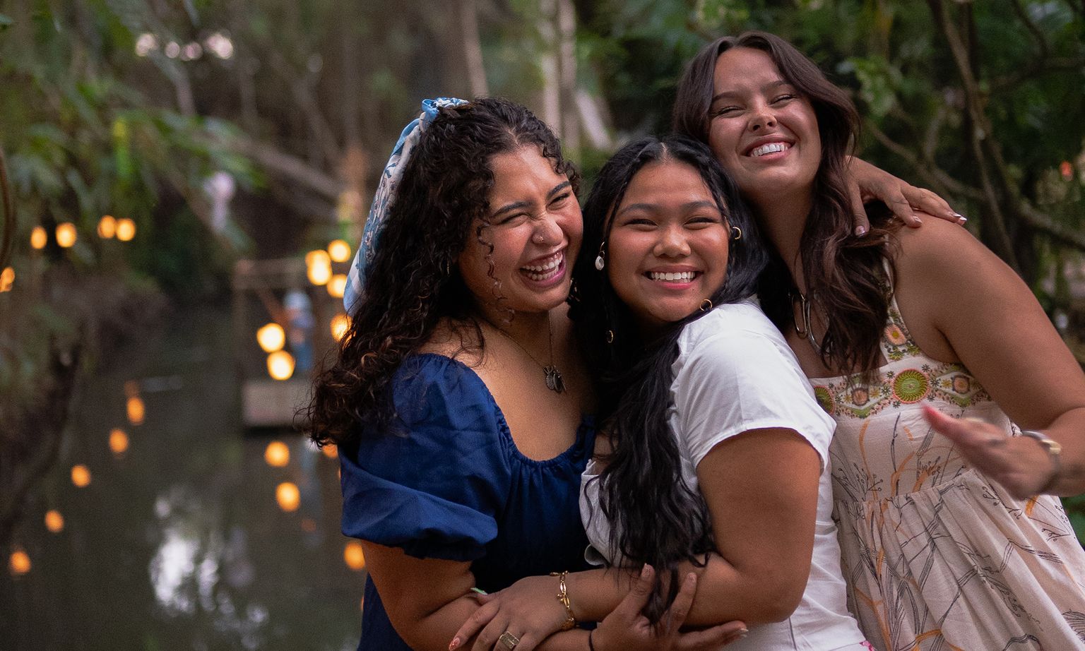 Three women happily embrace by a serene, lantern-lit riverside, surrounded by lush greenery.