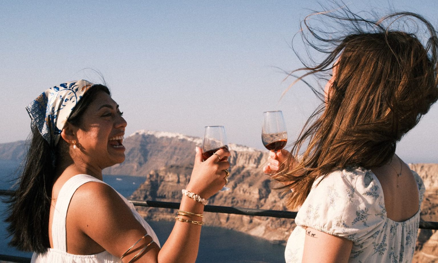 Two women laughing and clinking wine glasses on a balcony, with a scenic view of cliffs and the sea in the background.
