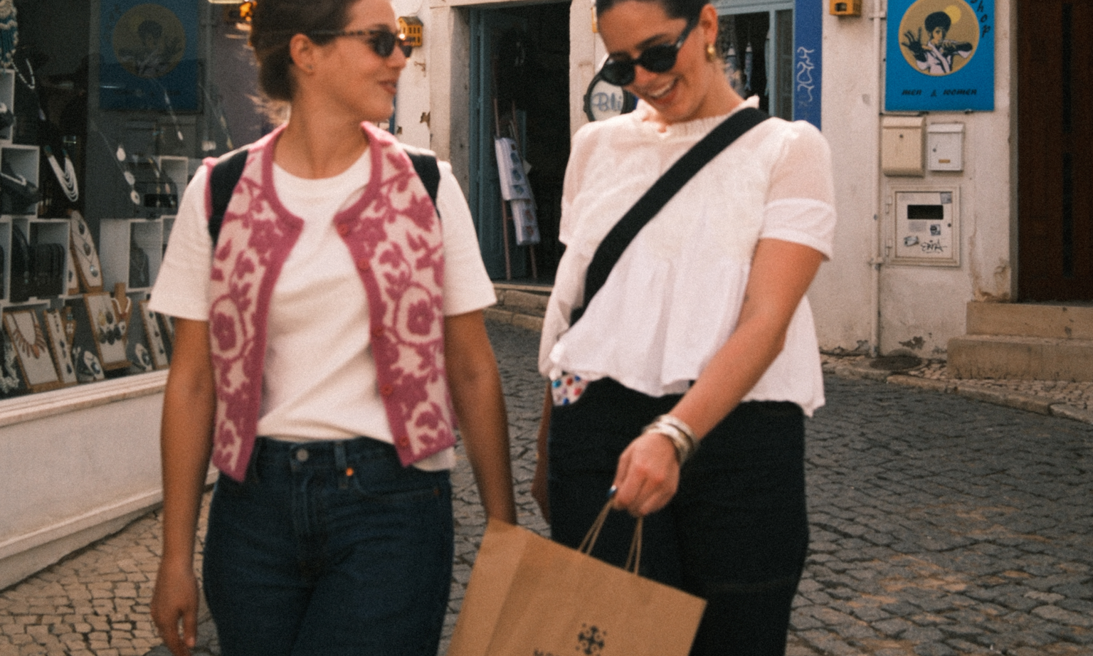 Two women wearing sunglasses, one holding a shopping bag, smiling in the streets of Portugal