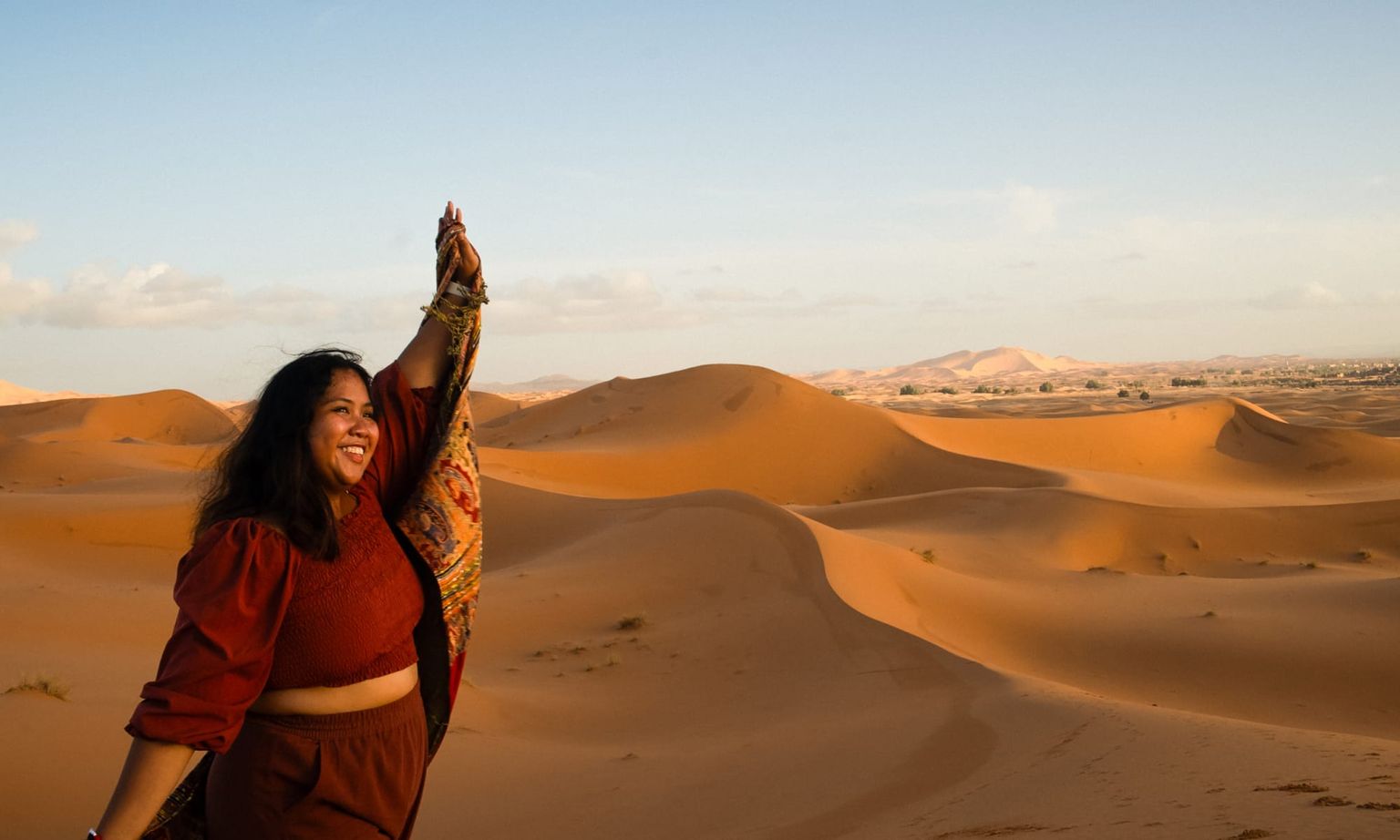 A woman joyfully holds a scarf in the air, standing in a vast desert landscape with rolling sand dunes under a clear blue sky.