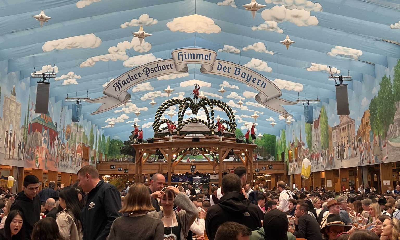 Inside the Hacker-Festzelt tent with a blue sky painted ceiling, clouds, and people walking around.