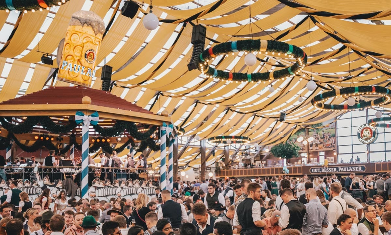 Inside the Paulaner Festzelt Oktoberfest tent with people sitting at tables, a band playing in the middle gazebo, and a giant "Paulaner" stein on top of the gazebo.