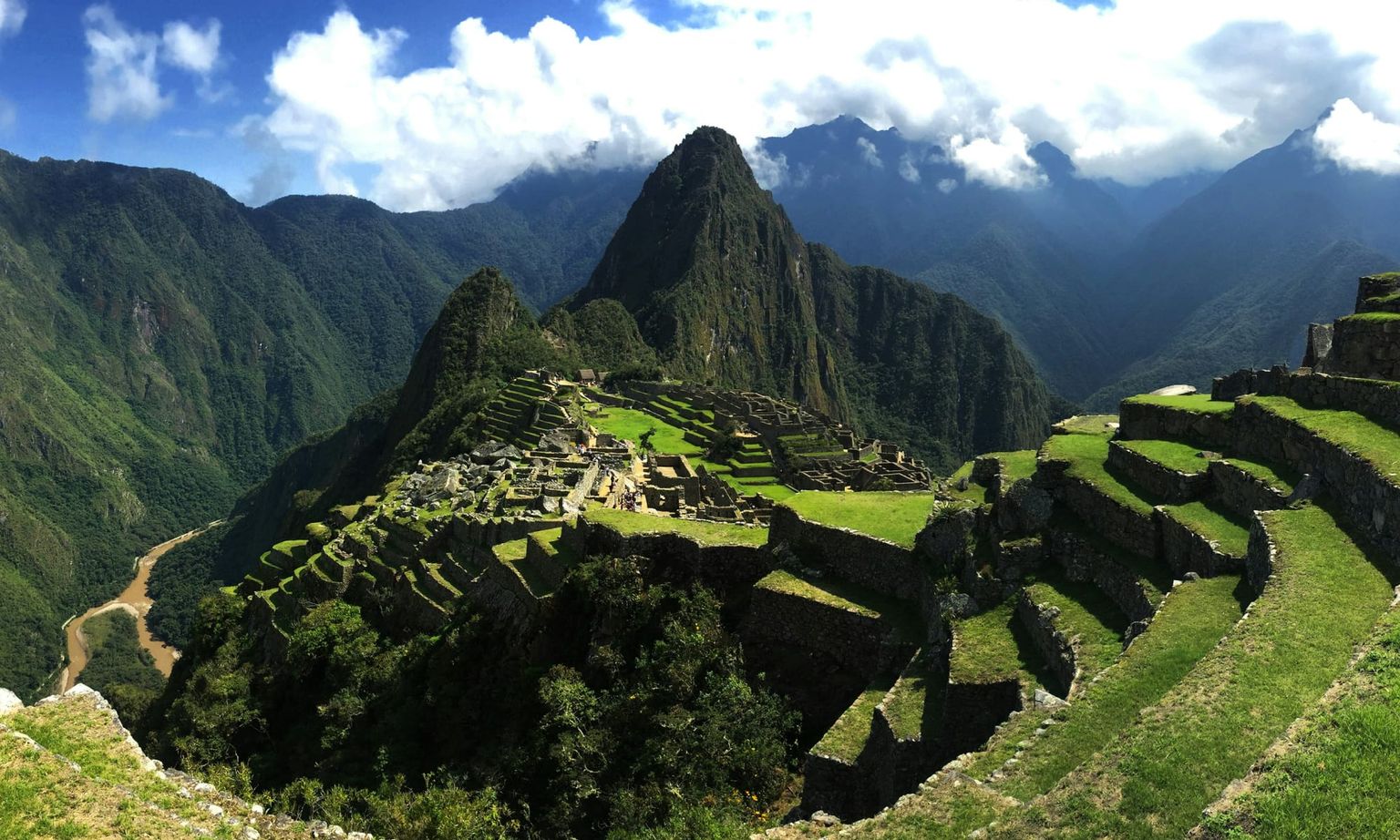 View of Machu Picchu with terraced green hills and rugged mountains under a partly cloudy sky.