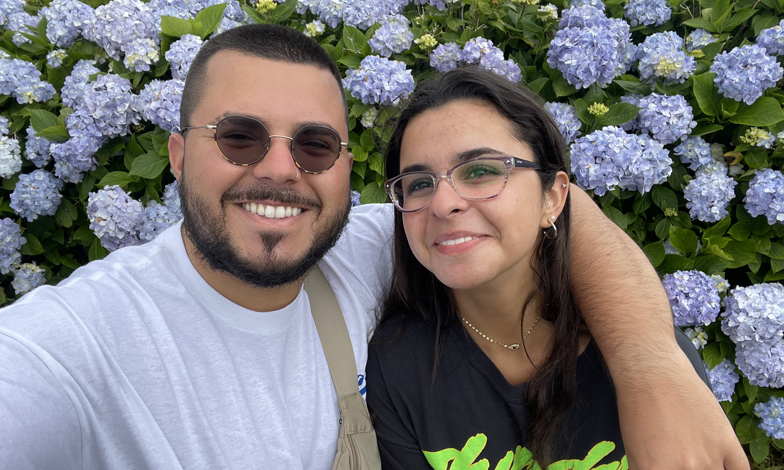 A man with arm around a woman, both smiling, in front of a blue hydrangea bush