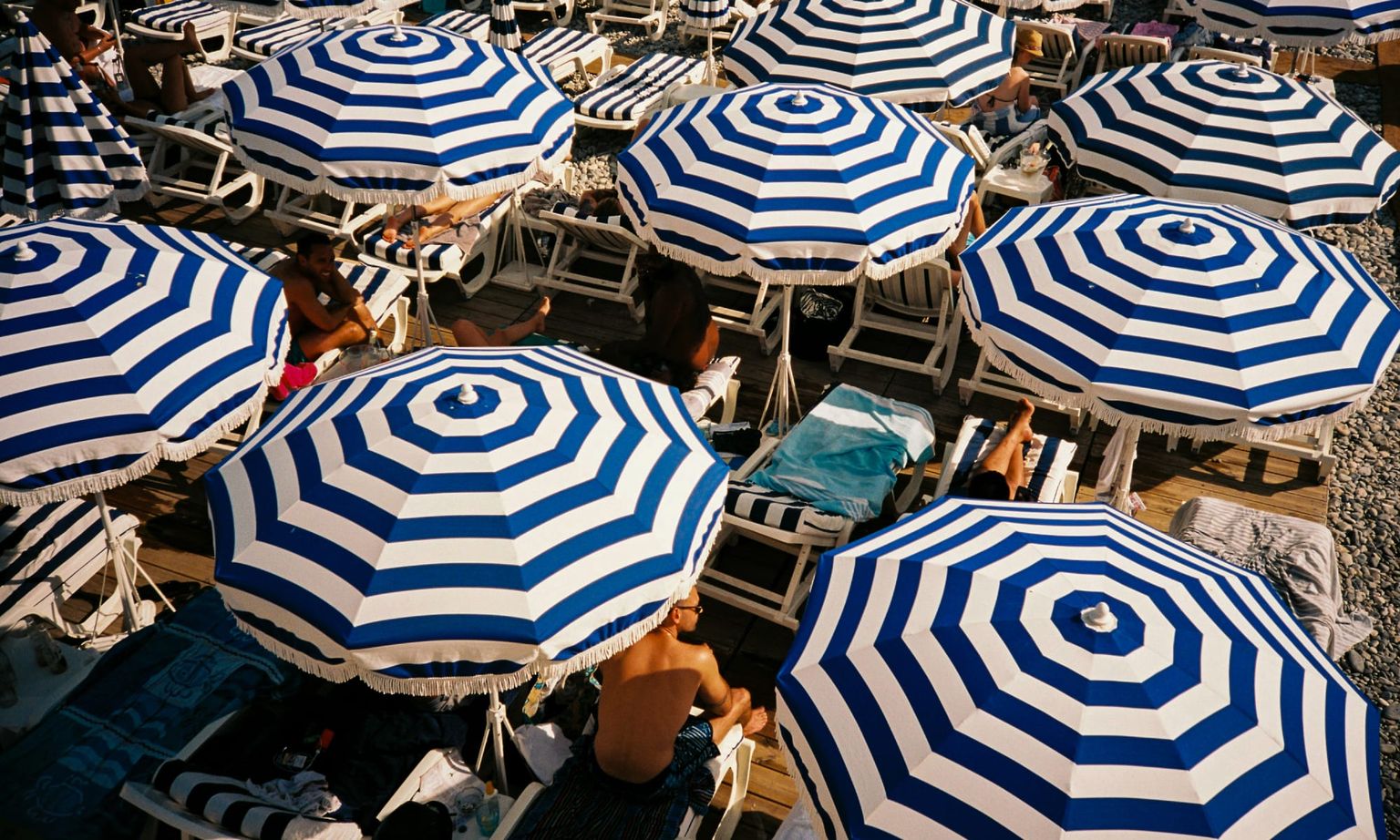 Blue and white striped umbrellas on a crowded pebble beach, with sunbathers resting on lounge chairs beneath them.