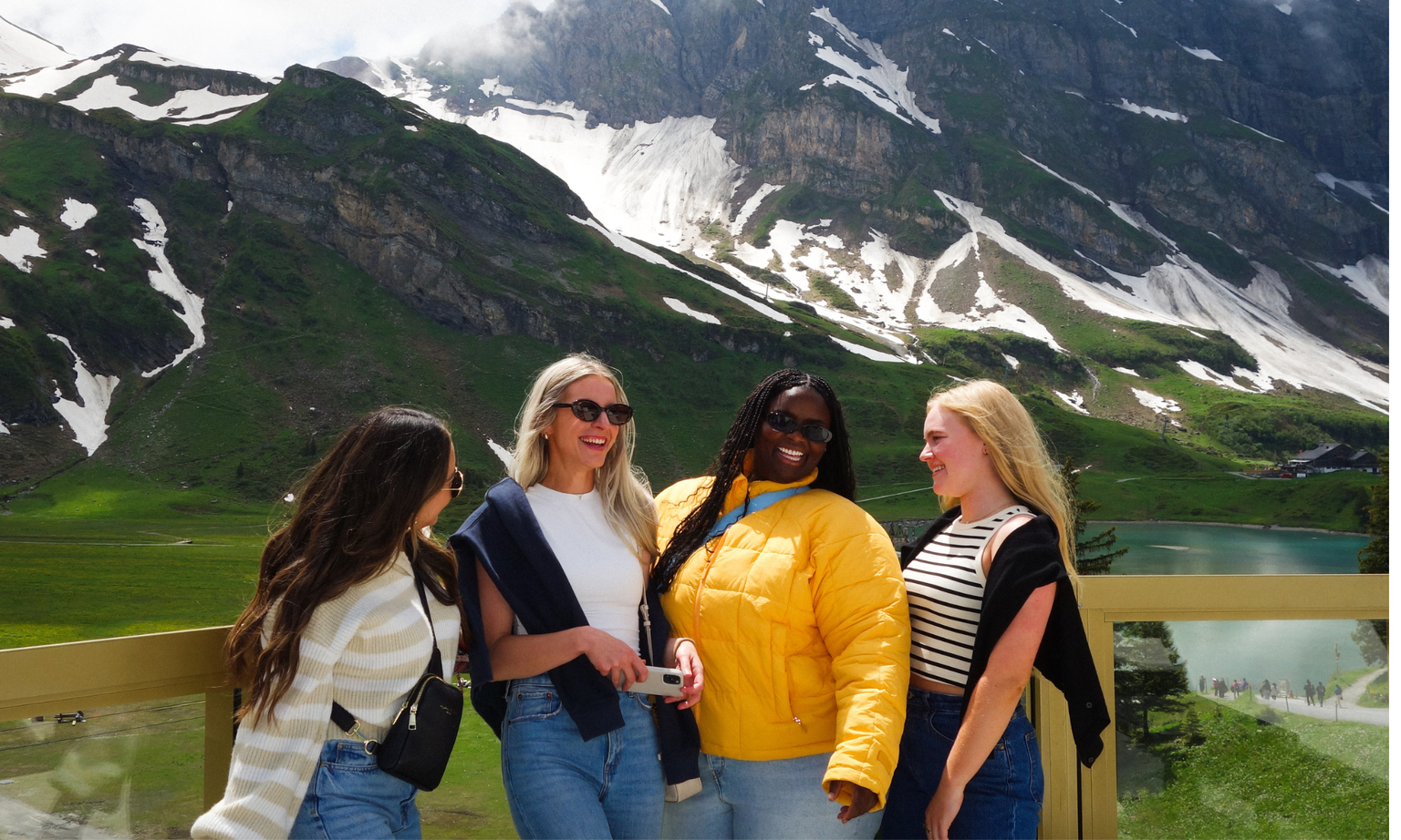 Four women smiling and posing with a scenic mountain landscape in the background, featuring snow patches and a lake.