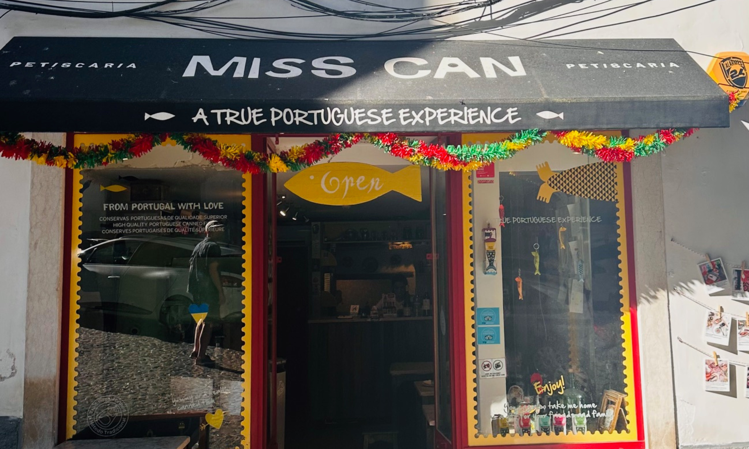 Front view of "Miss Can" shop with a black awning, colorful garlands, and signage reading "A True Portuguese Experience."