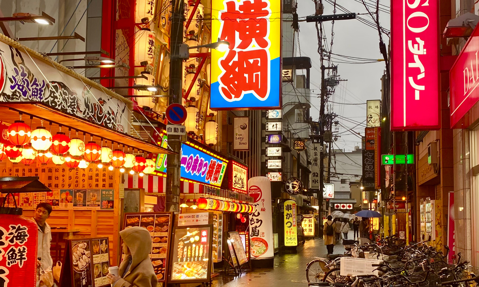Rainy urban street in Japan, lined with colorful neon signs and lanterns. People with umbrellas walk past restaurants and bicycles.