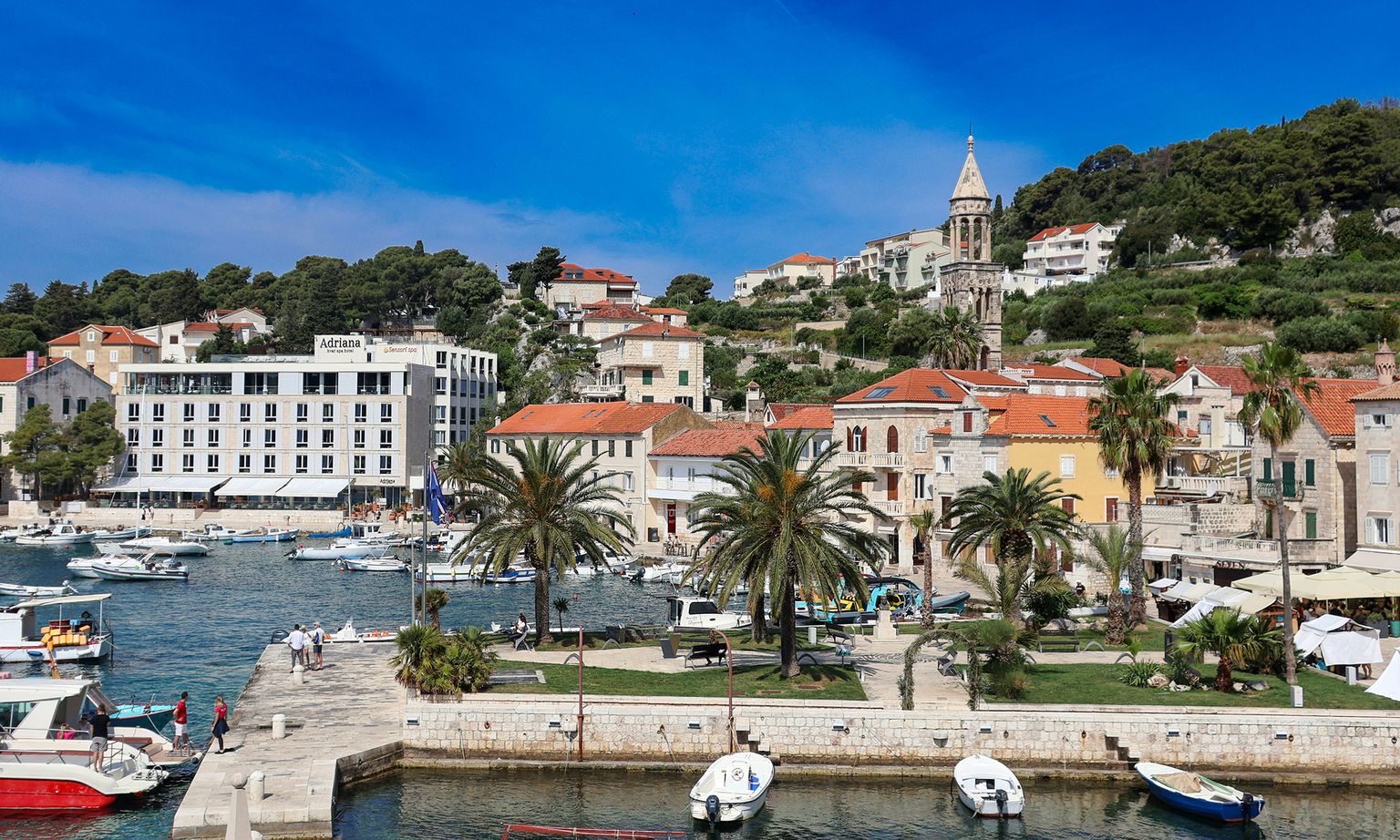Scenic coastal town with boats in a harbor, palm trees, colorful buildings, and a church steeple against a backdrop of lush hills under a blue sky.
