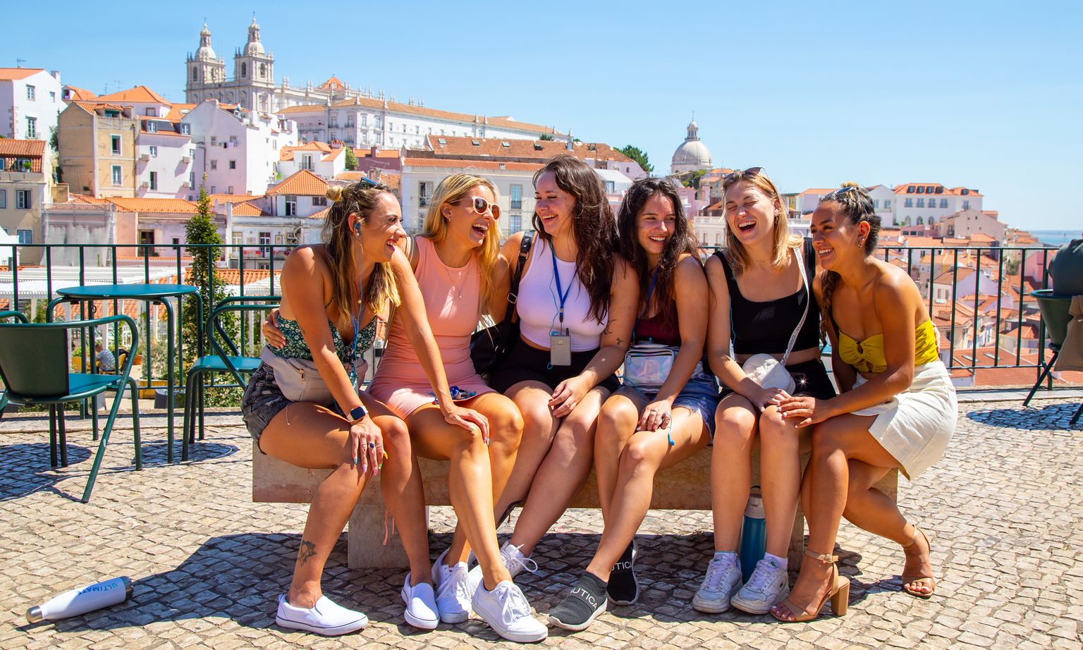 Six women sitting on a bench, laughing together, with a scenic view of a city and historical buildings in the background.