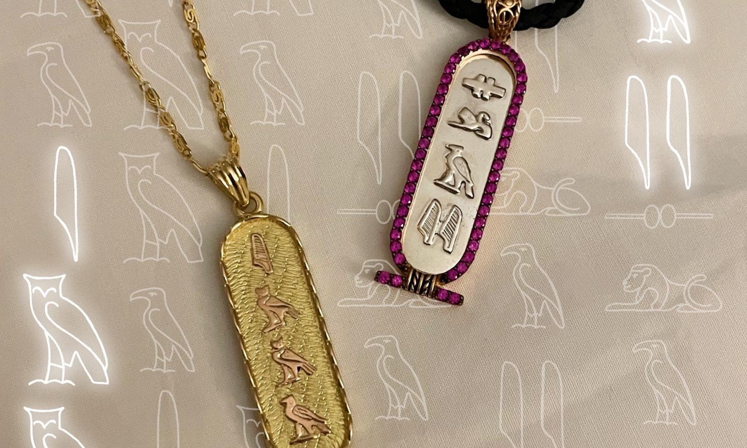 Two gold pendants with hieroglyphic designs, one bordered with pink stones, on a background with faint owl and cat outlines.