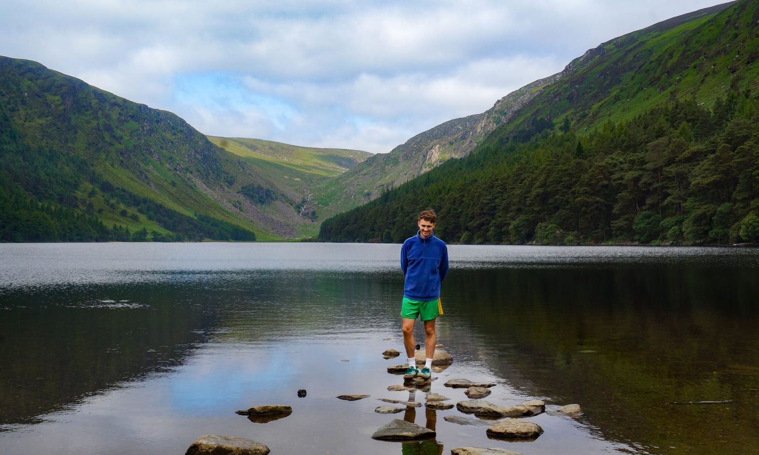 A person stands on stepping stones in a serene lake with surrounding green hills under a partly cloudy sky.