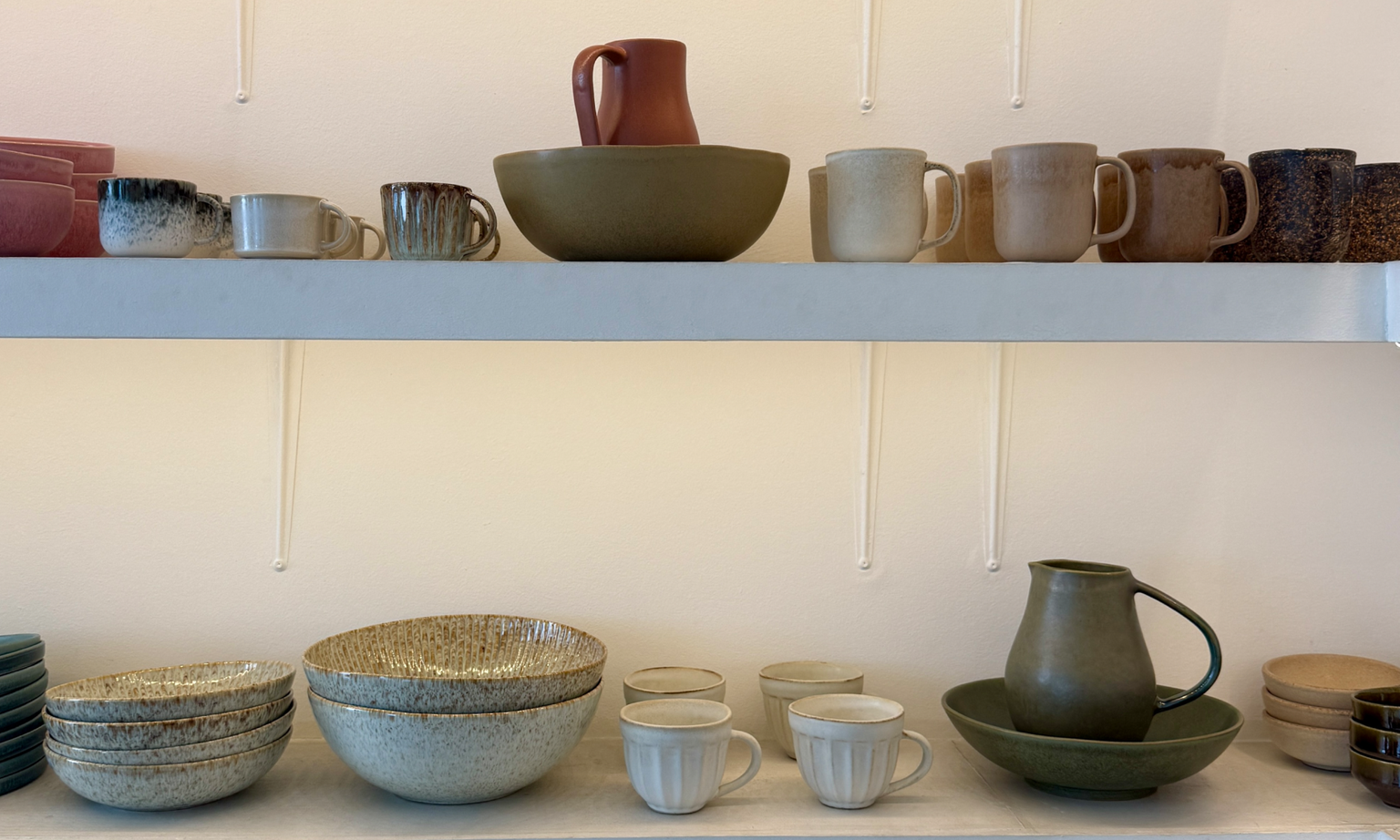 Two shelves of handmade ceramic mugs, cups, bowls, and pitchers