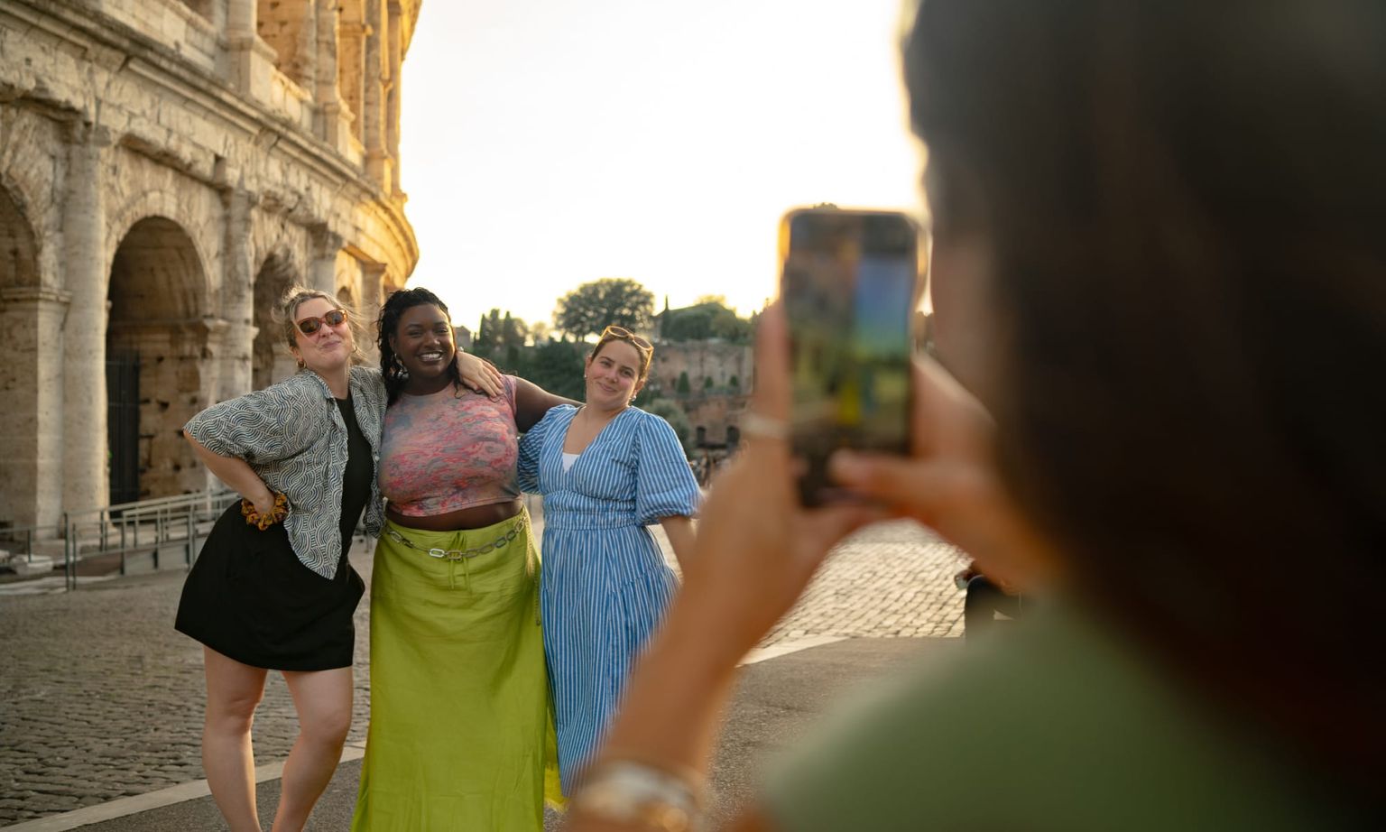 Three women pose for a photo in front of the Colosseum at sunset, with a person taking their picture using a smartphone in the foreground.