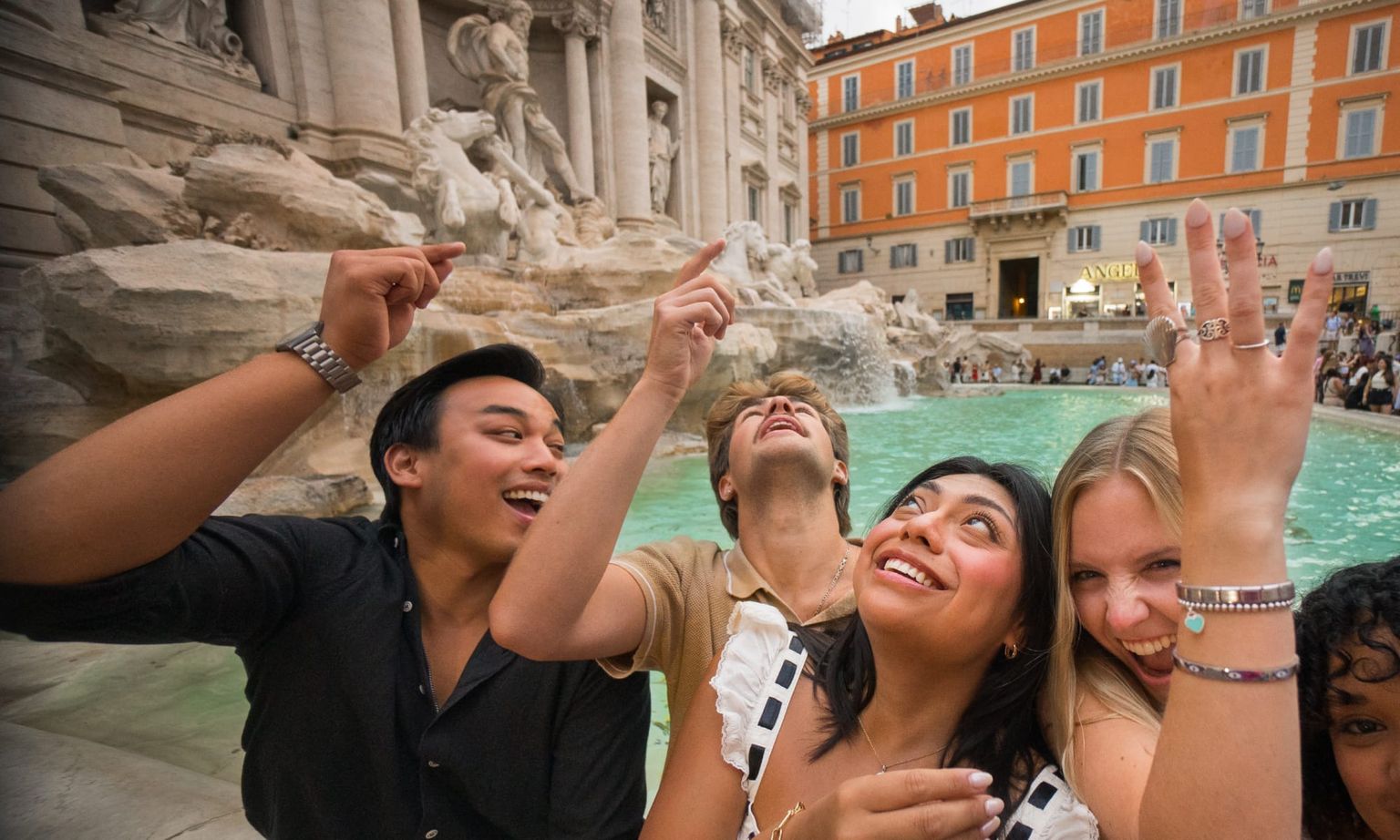 Four friends laugh and toss coins into the Trevi Fountain, surrounded by its ornate statues and a backdrop of colorful buildings.