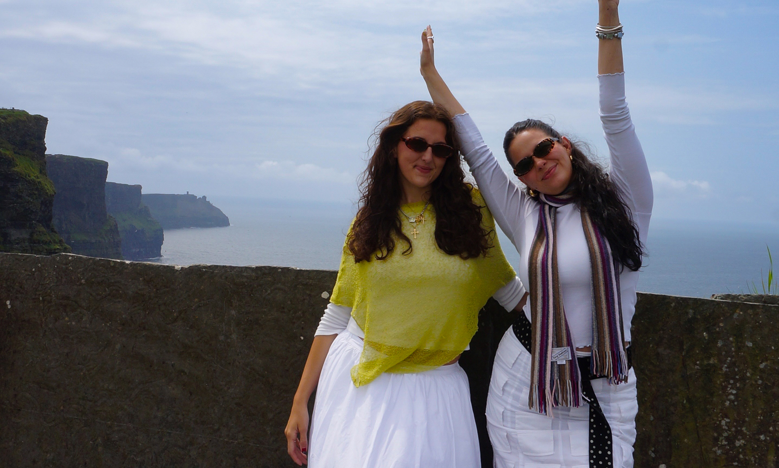 Two women pose happily by a stone wall near the Cliffs of Moher, Ireland, with the ocean in the background under a cloudy sky.