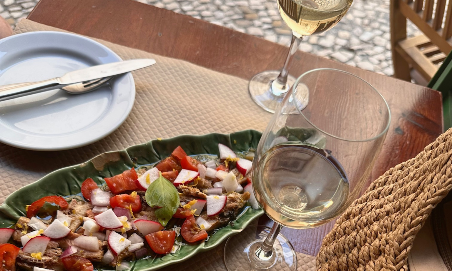 A green plate with a tomato and radish salad, two glasses of white wine, and a straw hat on a wooden table.