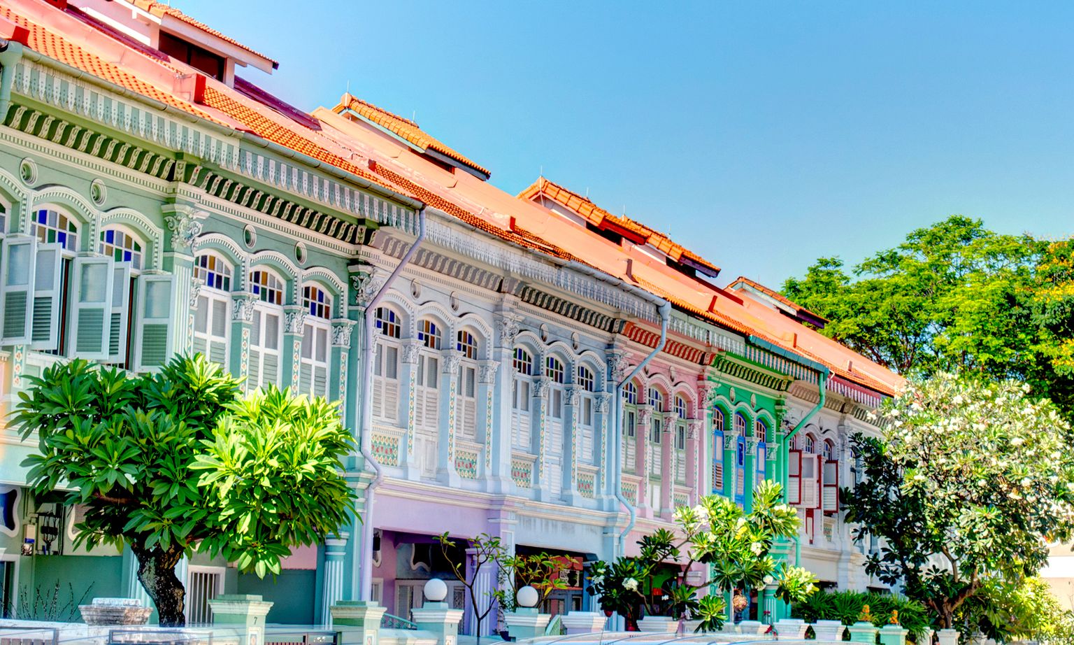 Colorful row of traditional shophouses with ornate pastel facades, red-tiled roofs, and greenery in front, under a clear blue sky.