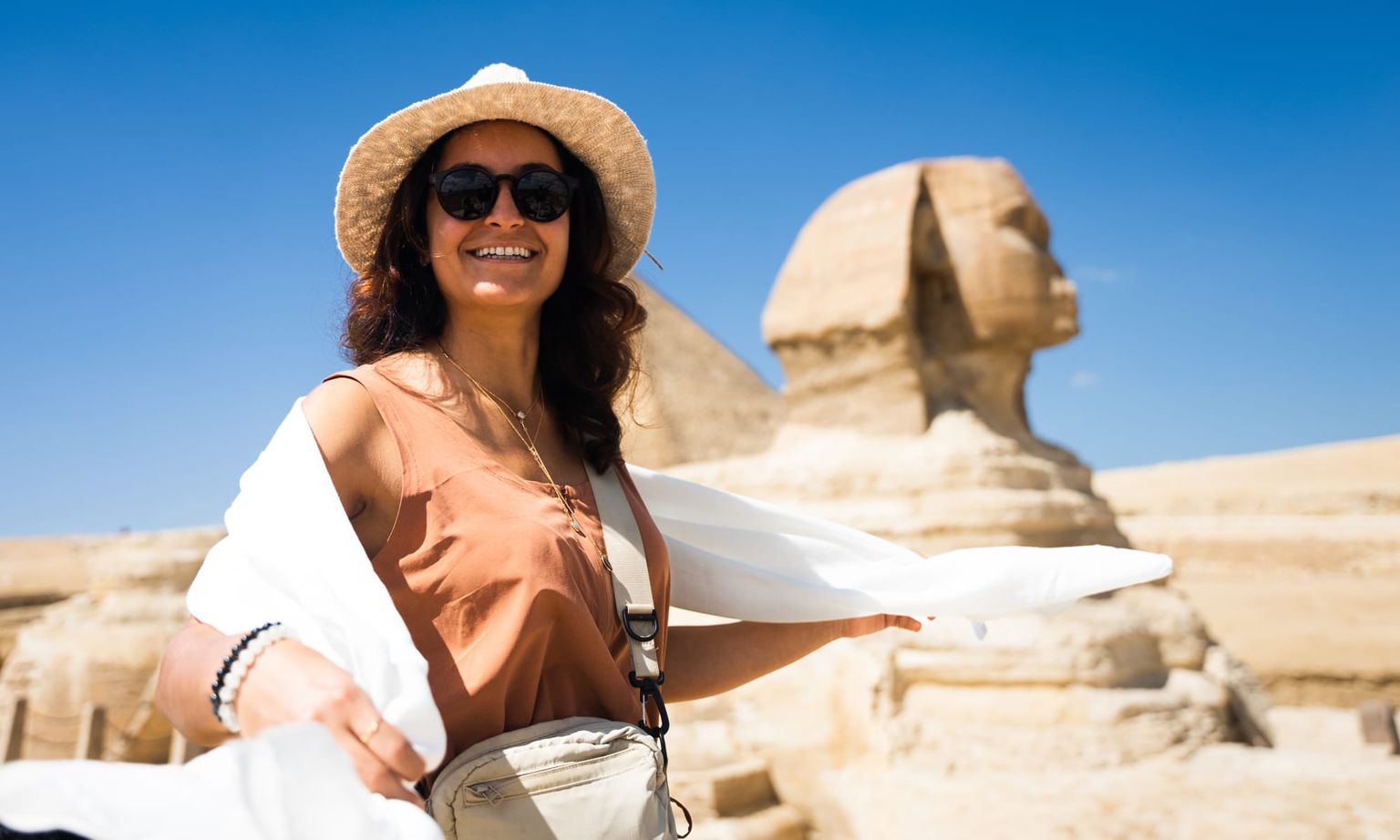 Woman in a sun hat and sunglasses poses happily in front of the Sphinx under a clear blue sky.