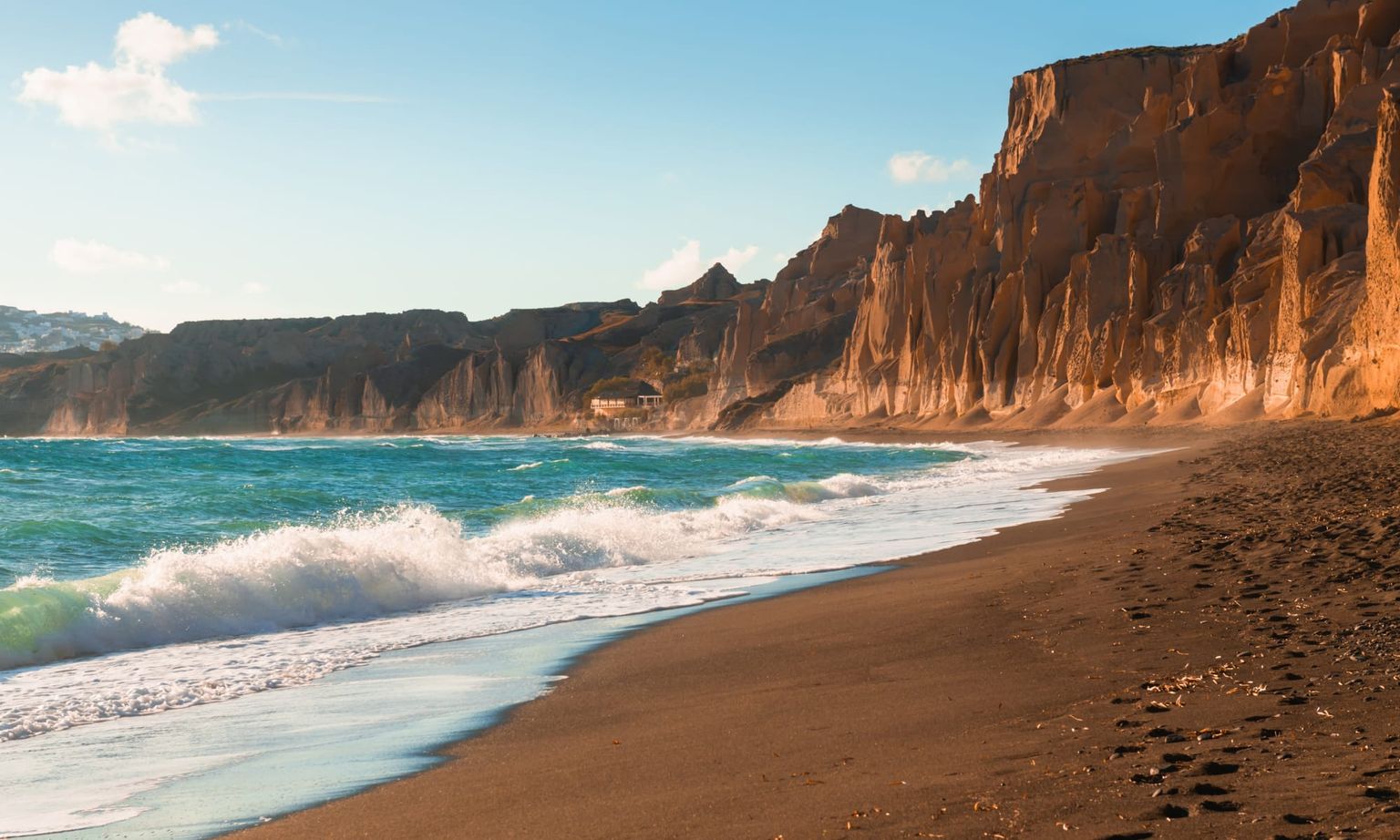 Waves crashing on a sandy beach with towering, jagged cliffs under a clear blue sky. A small building is visible in the distance.