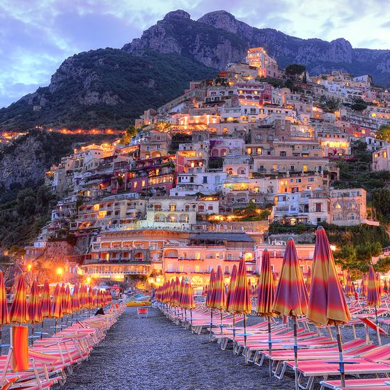 A picturesque coastal village with colorful buildings on a hillside, lit up at dusk. Rows of orange beach umbrellas line the foreground.