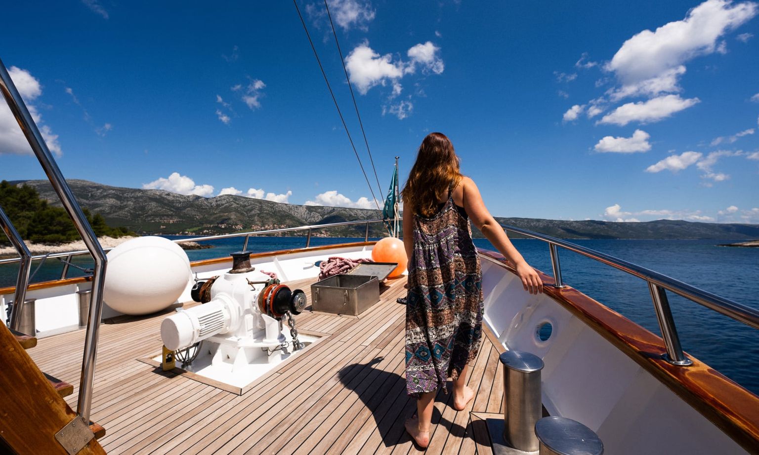Woman in a patterned dress stands on a boat deck, gazing at mountainous shoreline under a clear blue sky.