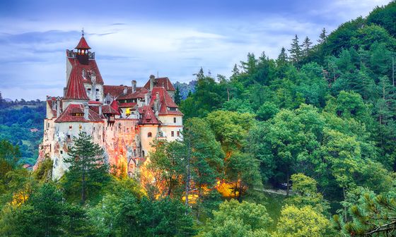 A gothic-style castle with red roofs, surrounded by lush green forest under a cloudy sky, illuminated by warm lights in the evening.