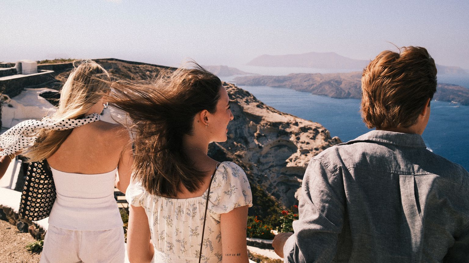 Three people facing the sea, with wind blowing their hair, overlooking a scenic coastal view with cliffs and clear blue water under a bright sky.