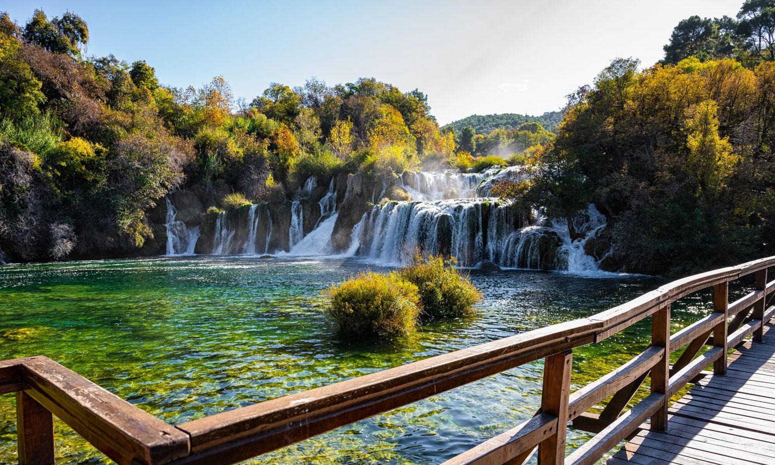 Wooden boardwalk beside a clear, green river leading to small cascading waterfalls, surrounded by lush trees under a partly cloudy sky.