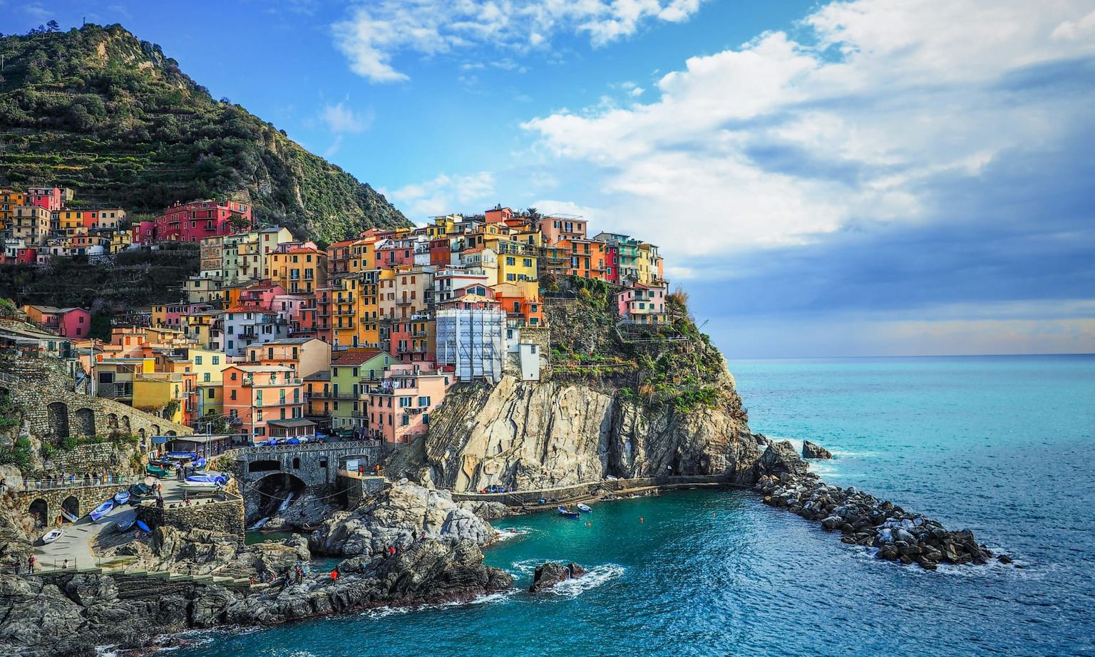Colorful buildings perched on a rocky cliff above a turquoise sea, with a mountain backdrop and partly cloudy sky in Cinque Terre, Italy.