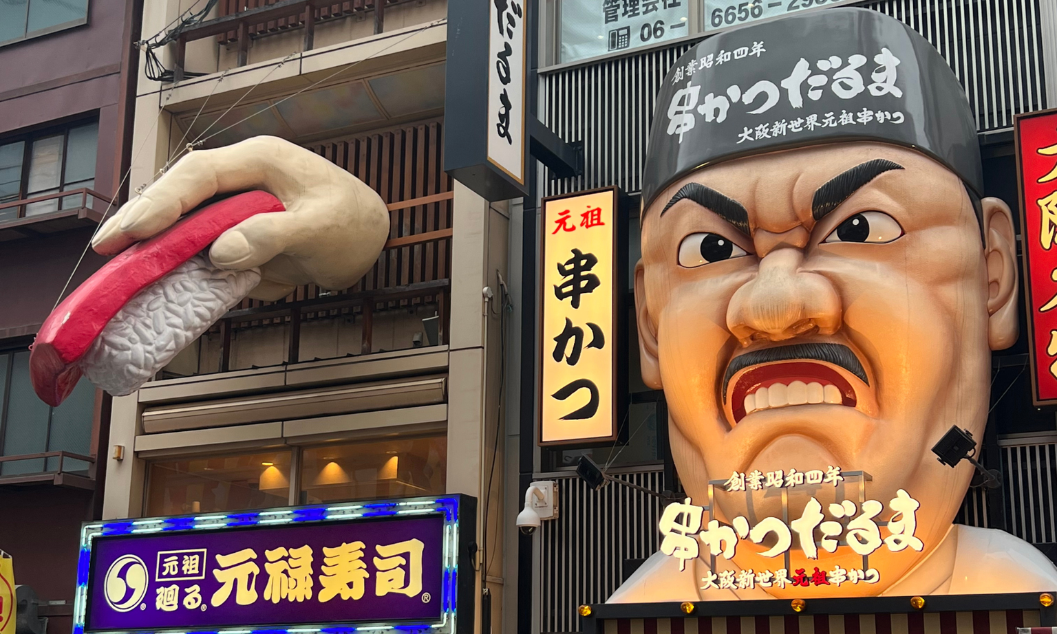 Colorful storefronts in Osaka featuring a giant sushi hand and a large, expressive face sculpture on building facades.