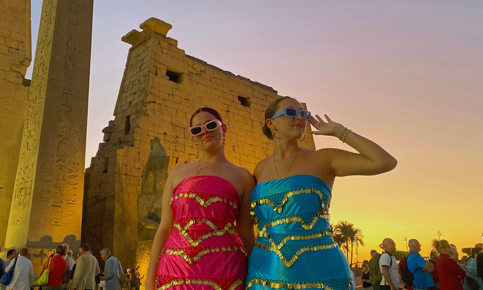 Two women in vibrant dresses and sunglasses pose in front of ancient ruins at sunset, with a crowd and palm trees in the background.