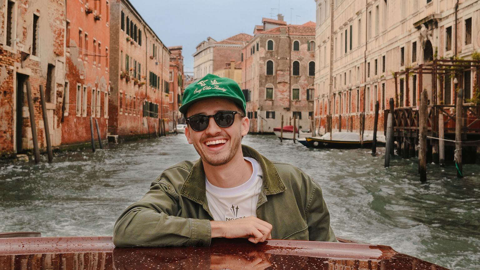 A smiling person wearing sunglasses and a green cap leans on a boat, cruising through a canal lined with historic buildings.