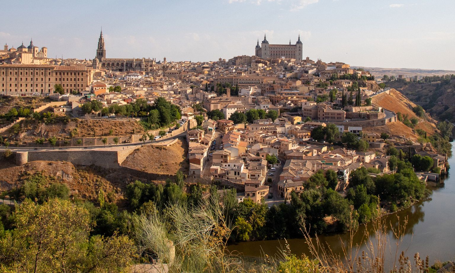 Aerial view of Toledo, Spain, showing historic buildings, a cathedral, greenery, and the Tagus River under a clear sky.