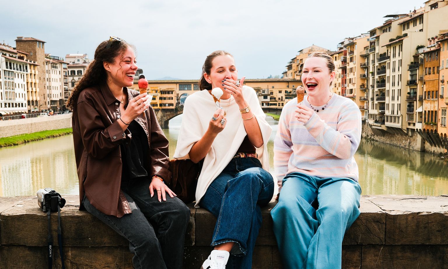 Three people sitting on a stone ledge, laughing and eating ice cream cones near a river with historic buildings in the background.