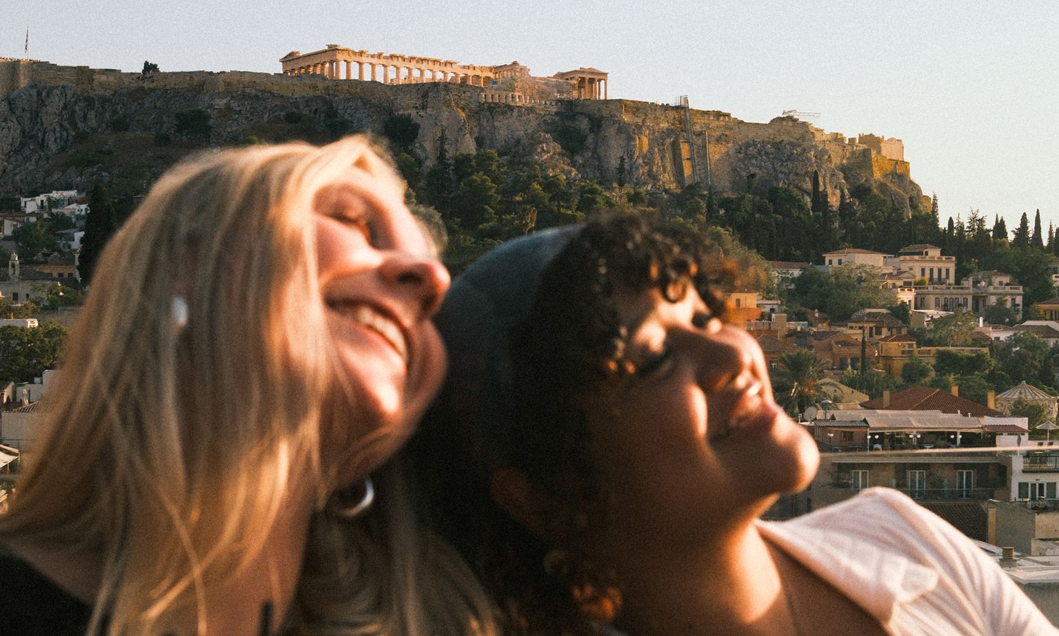 Two smiling people with their heads together, with the Acropolis and Parthenon in the background at sunset.
