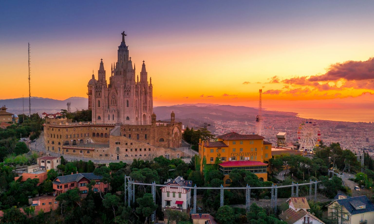 Aerial view of the Temple Expiatori del Sagrat Cor on a hill at sunset, overlooking a cityscape and ocean with a Ferris wheel visible.